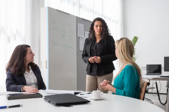 Three women in an office setting having a professional discussion, focused and engaged with a whiteboard. How to define a lead generation software target audience